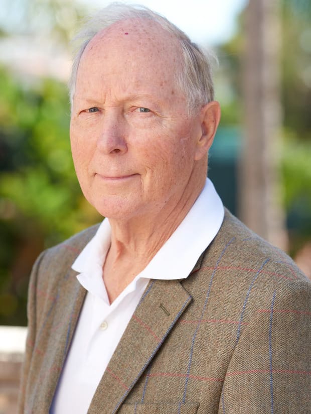 Elderly man with a calm expression, wearing a gray plaid blazer and white shirt. He stands outdoors with blurred greenery in the background.