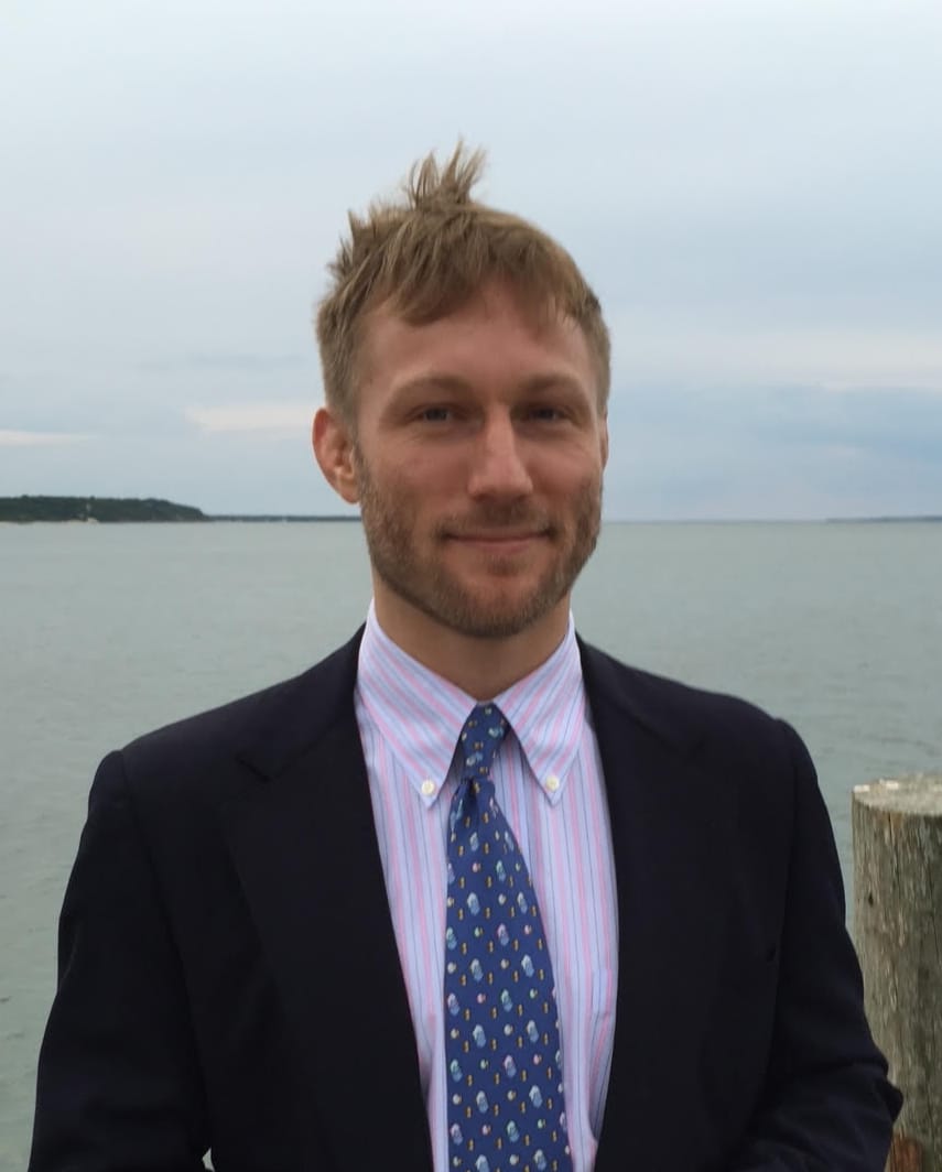 A man in a suit and tie stands by a calm, wide body of water with a cloudy sky. His hair is slightly windswept, and he shows a subtle smile.