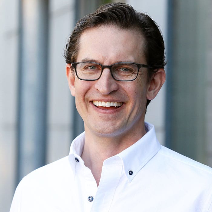 A man with glasses smiles warmly, wearing a white button-up shirt. He stands against a blurred background of tall building columns, conveying a professional yet approachable demeanor.