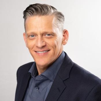 A smiling man in a dark blazer and blue shirt stands against a light gray background. His confident expression conveys a professional and approachable tone.