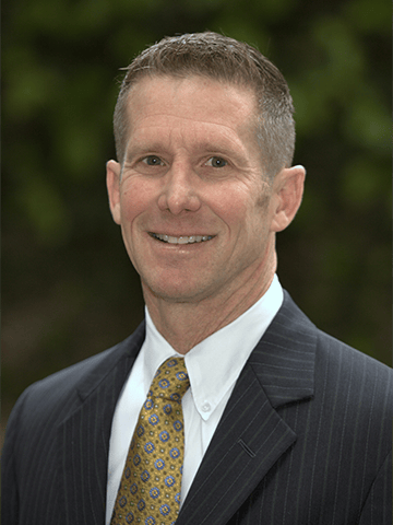 A man in a pinstripe suit, white shirt, and patterned tie poses outdoors in front of a blurred green background.