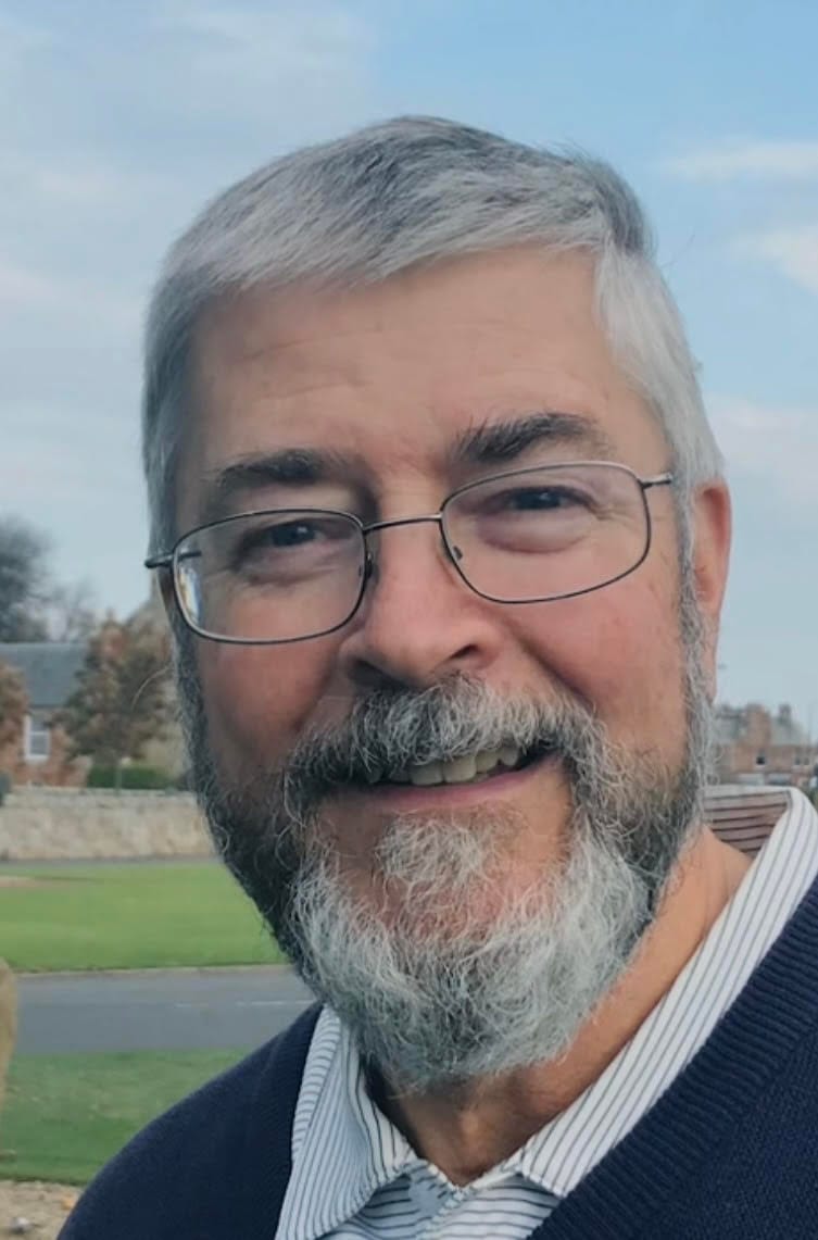 Elderly man with a gray beard and glasses smiles outdoors, standing against a backdrop of grass and houses under a clear blue sky, conveying warmth.